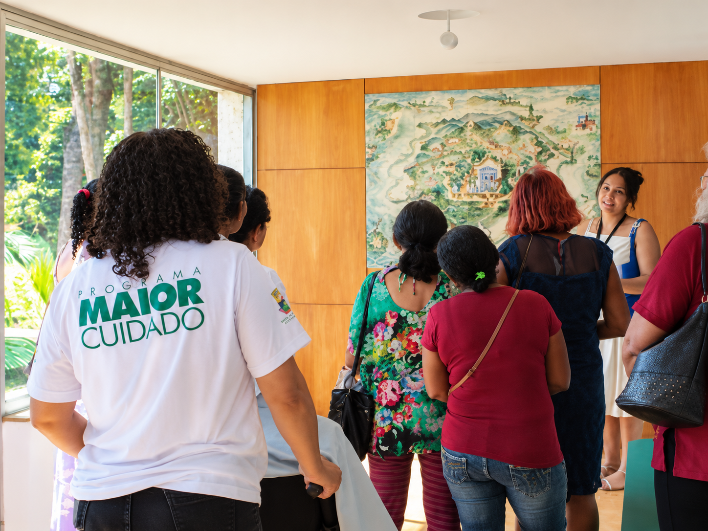 Grupo de mulheres, uma delas com camiseta do Programa Maior Cuidado, observando um painel decorativo em um ambiente interno