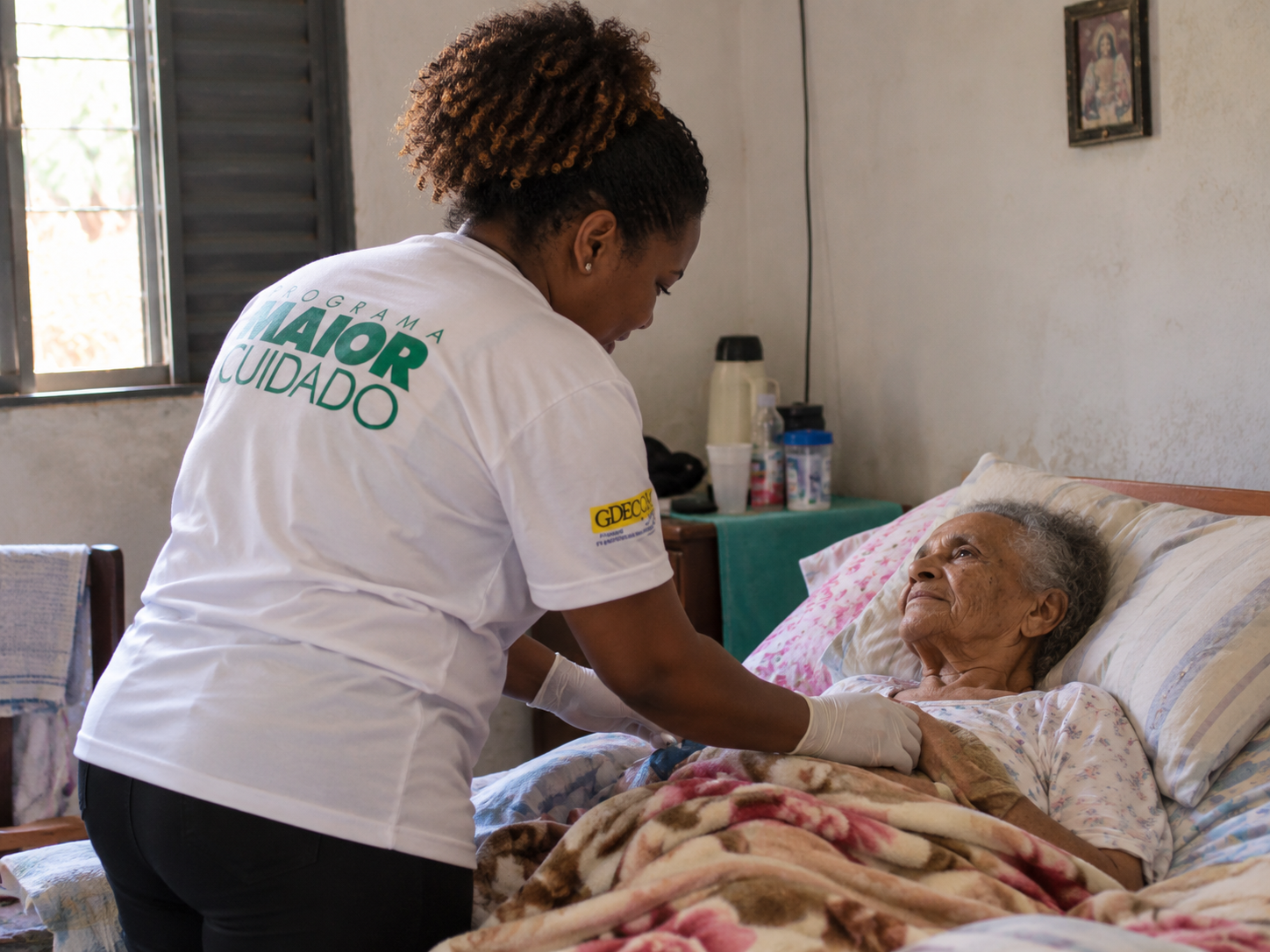Profissional do Programa Maior Cuidado prestando atendimento domiciliar a uma pessoa idosa, com as mãos apoiadas gentilmente sobre ela