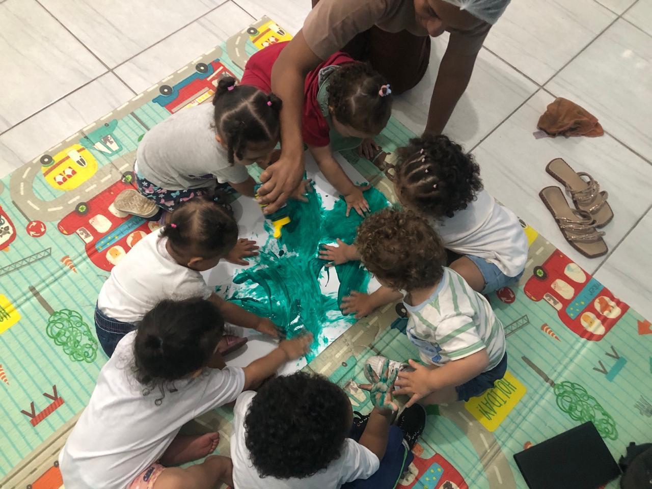 Crianças da Creche Patati-Patatá pintando coletivamente com tinta guache sobre papel no chão, acompanhadas por uma educadora