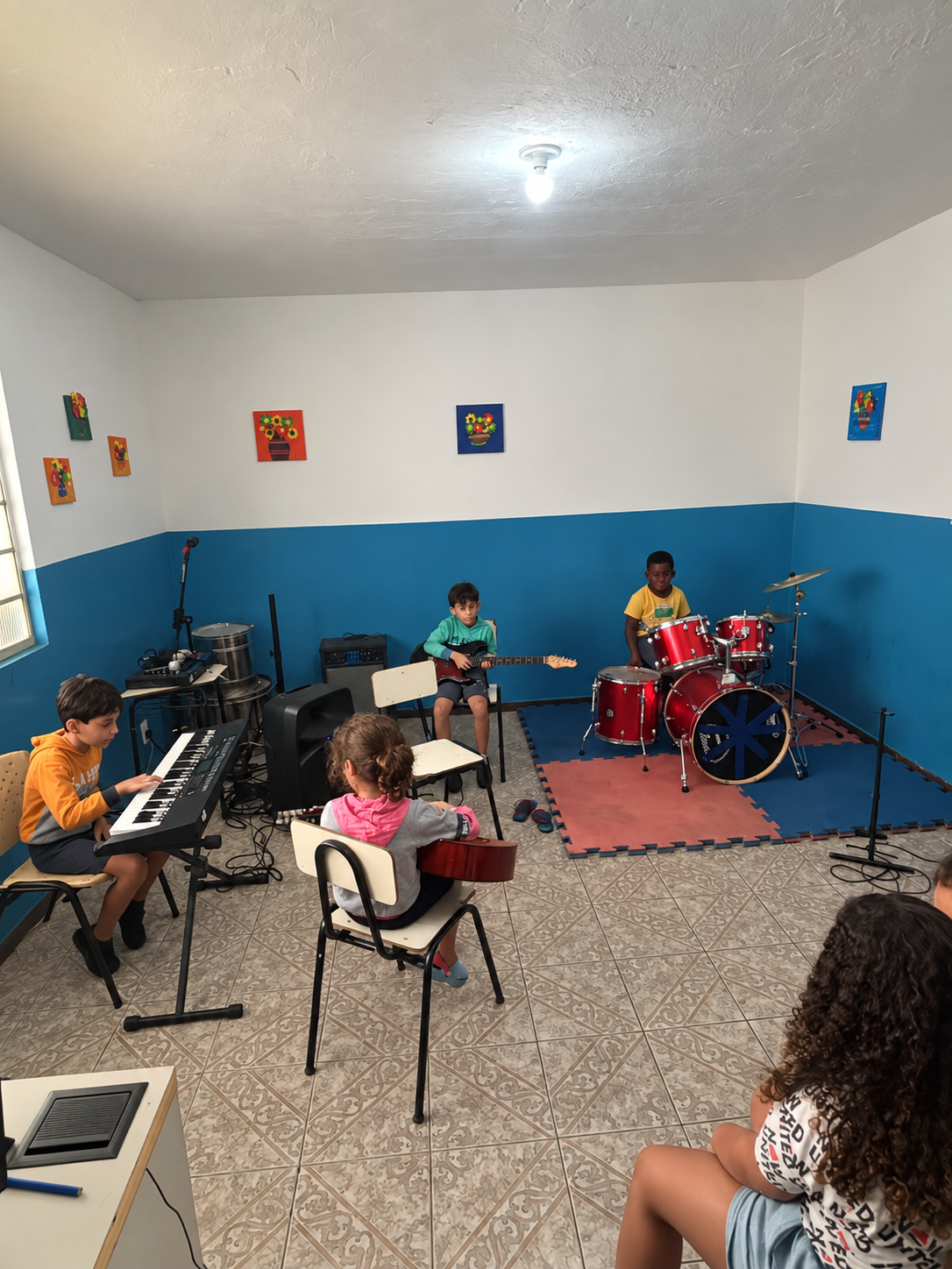 Crianças tocando teclado, violão e bateria em sala de música decorada com quadros coloridos