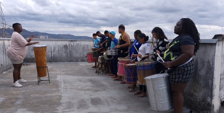Grupo de adolescentes do SCFV tocando instrumentos de percussão em fileira, ao ar livre, com uma educadora à frente